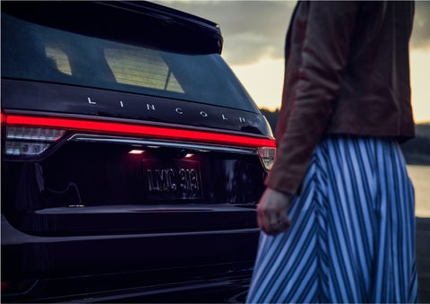 A person is shown near the rear of a 2023 Lincoln Aviator® SUV as the Lincoln Embrace illuminates the rear lights | Vance Lincoln in Miami OK