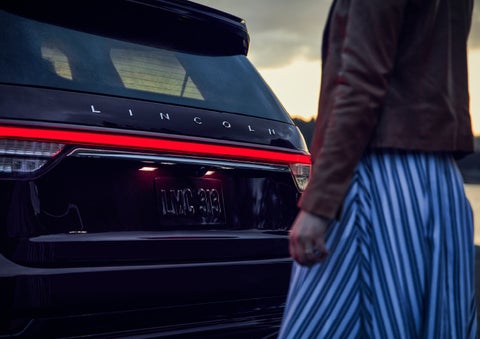 A person is shown near the rear of a 2024 Lincoln Aviator® SUV as the Lincoln Embrace illuminates the rear lights | Vance Lincoln in Miami OK