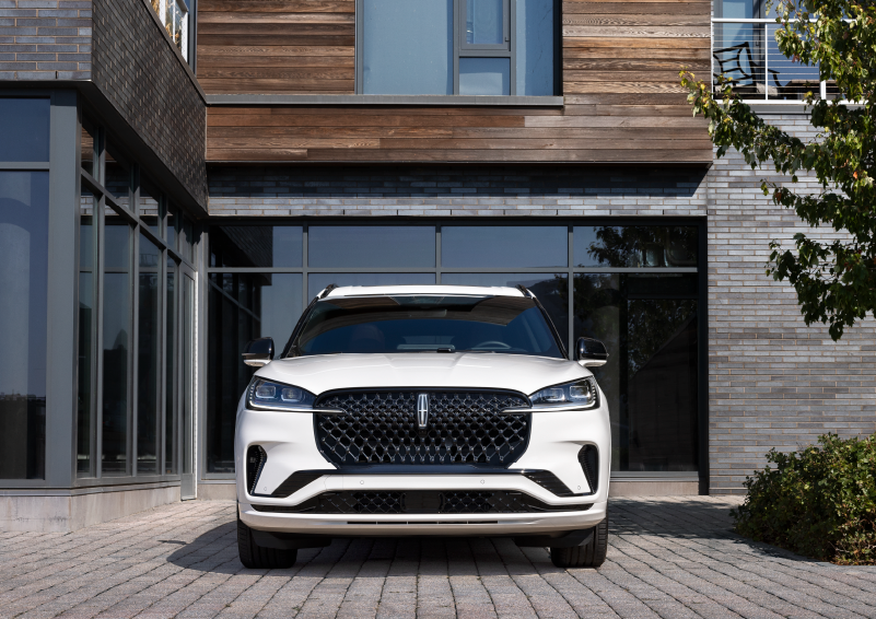 A white 2025 Lincoln Aviator® SUV is shown parked outside a modern building with gray brick and wood accents. | Vance Lincoln in Miami OK