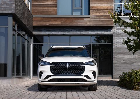 A white 2025 Lincoln Aviator® SUV is shown parked outside a modern building with gray brick and wood accents. | Vance Lincoln in Miami OK