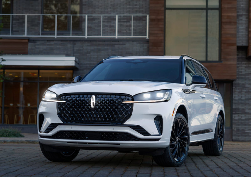 A white 2025 Lincoln Aviator® SUV is shown parked outside a modern building with gray brick and wood accents. | Vance Lincoln in Miami OK