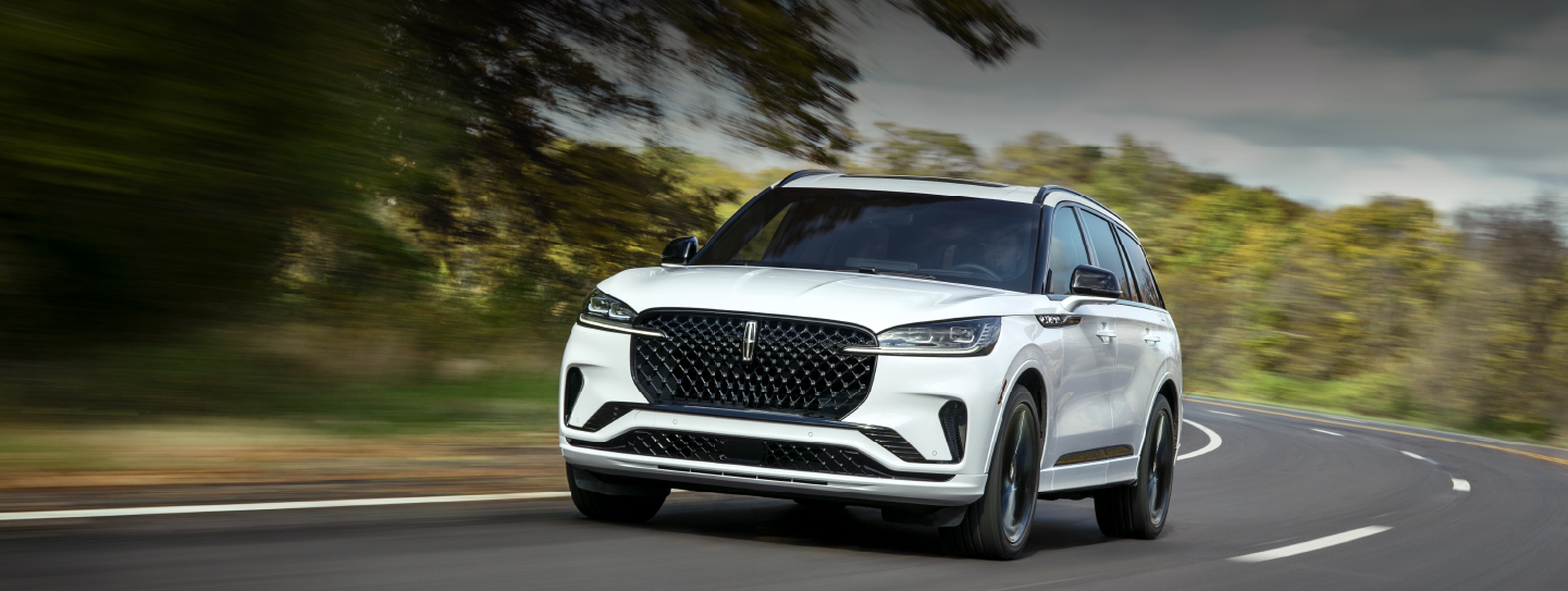 A white 2025 Lincoln Aviator® SUV is being driven down a road lined with trees. | Vance Lincoln in Miami OK