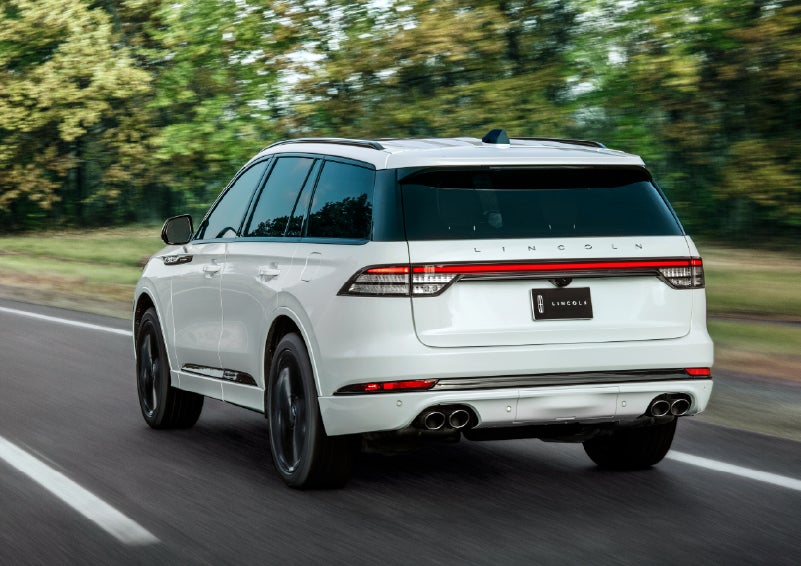 A white 2026 Lincoln Aviator® SUV is being driven away down a road lined with colorful trees. | Vance Lincoln in Miami OK