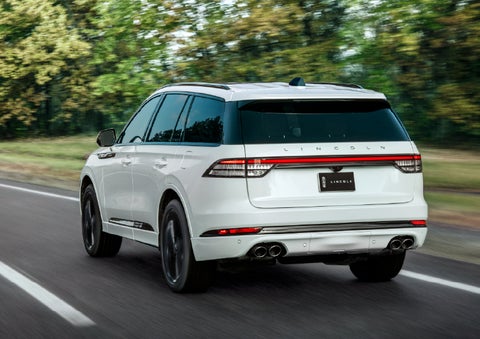 A white 2026 Lincoln Aviator® SUV is being driven away down a road lined with colorful trees. | Vance Lincoln in Miami OK